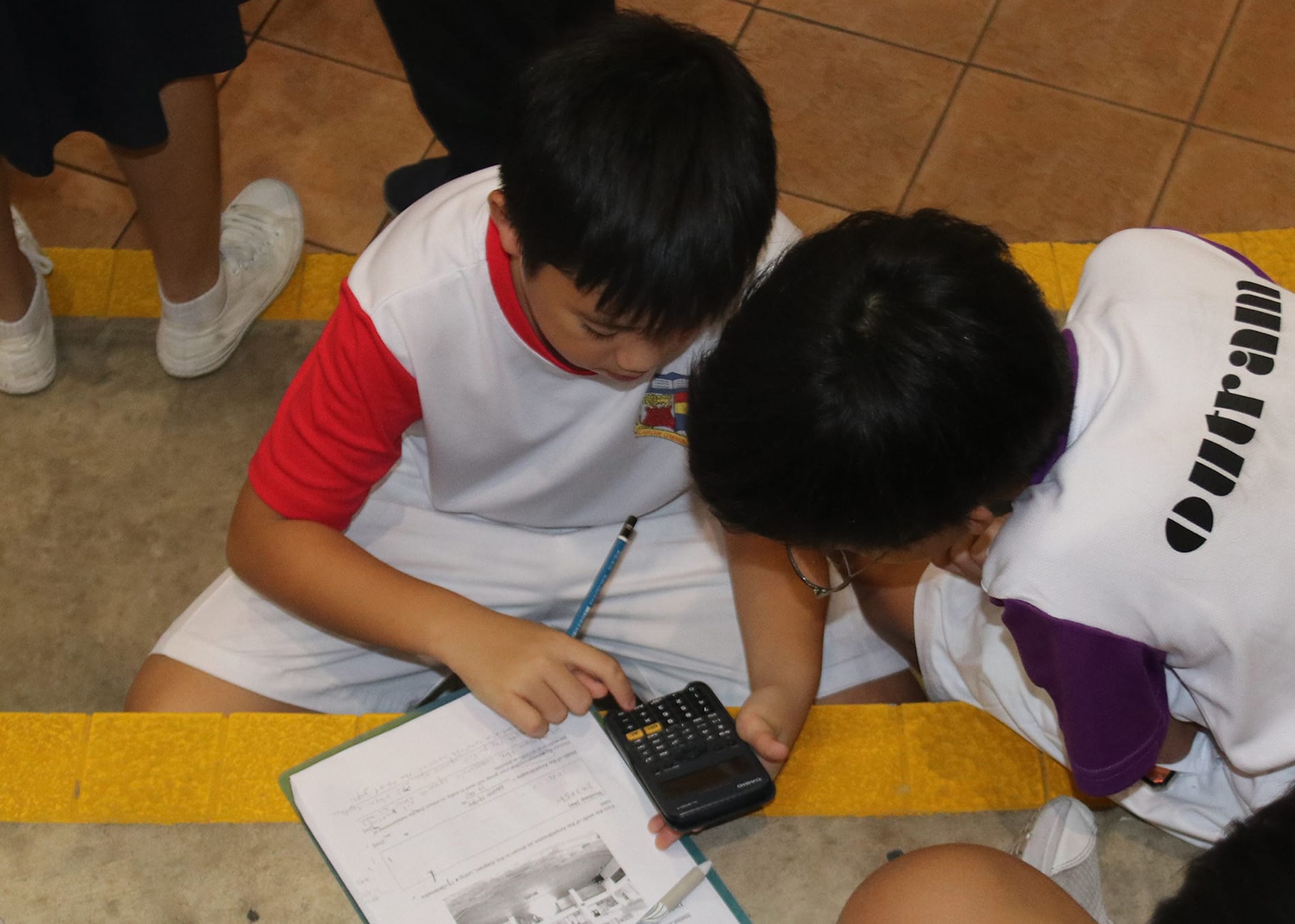 Two school boys in uniforms sit on a tiled floor, looking at a calculator and a worksheet together.
